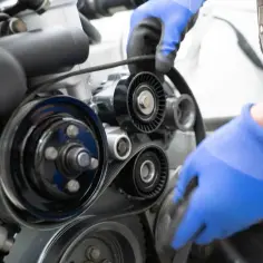 Mechanik w niebieskich rękawiczkach wymienia pasek rozrządu w Volvo V50. Widoczne koła pasowe i napinacz.