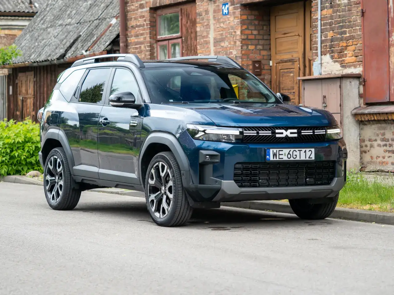 Niebieski SUV Dacia Duster z polskimi tablicami WE 6GT12 zaparkowany przed ceglanym budynkiem.