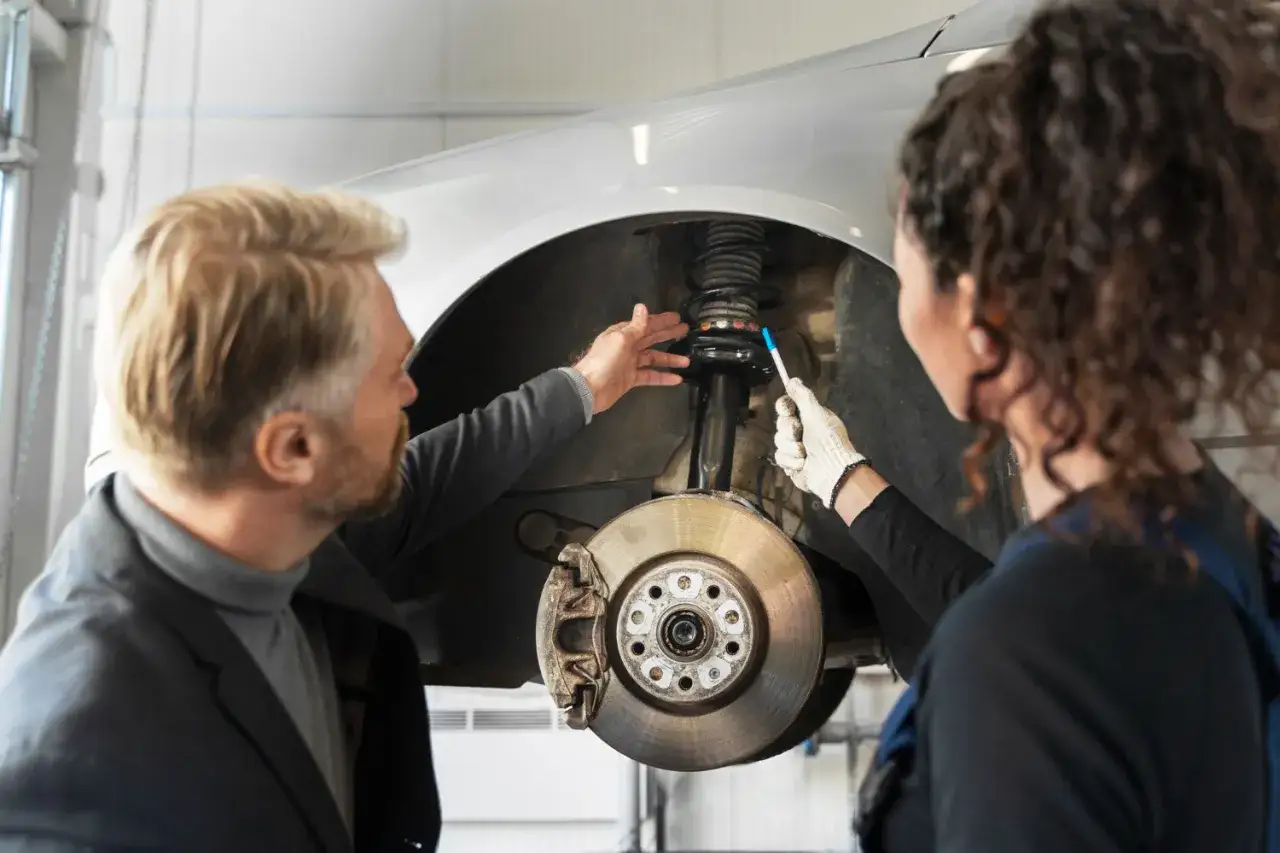 Mechanik i klientka dyskutują o amortyzatorach. Klientka pyta, ile wytrzymuje dwumasa w 2.0 TDI, gdy mechanik wskazuje na element zawieszenia.