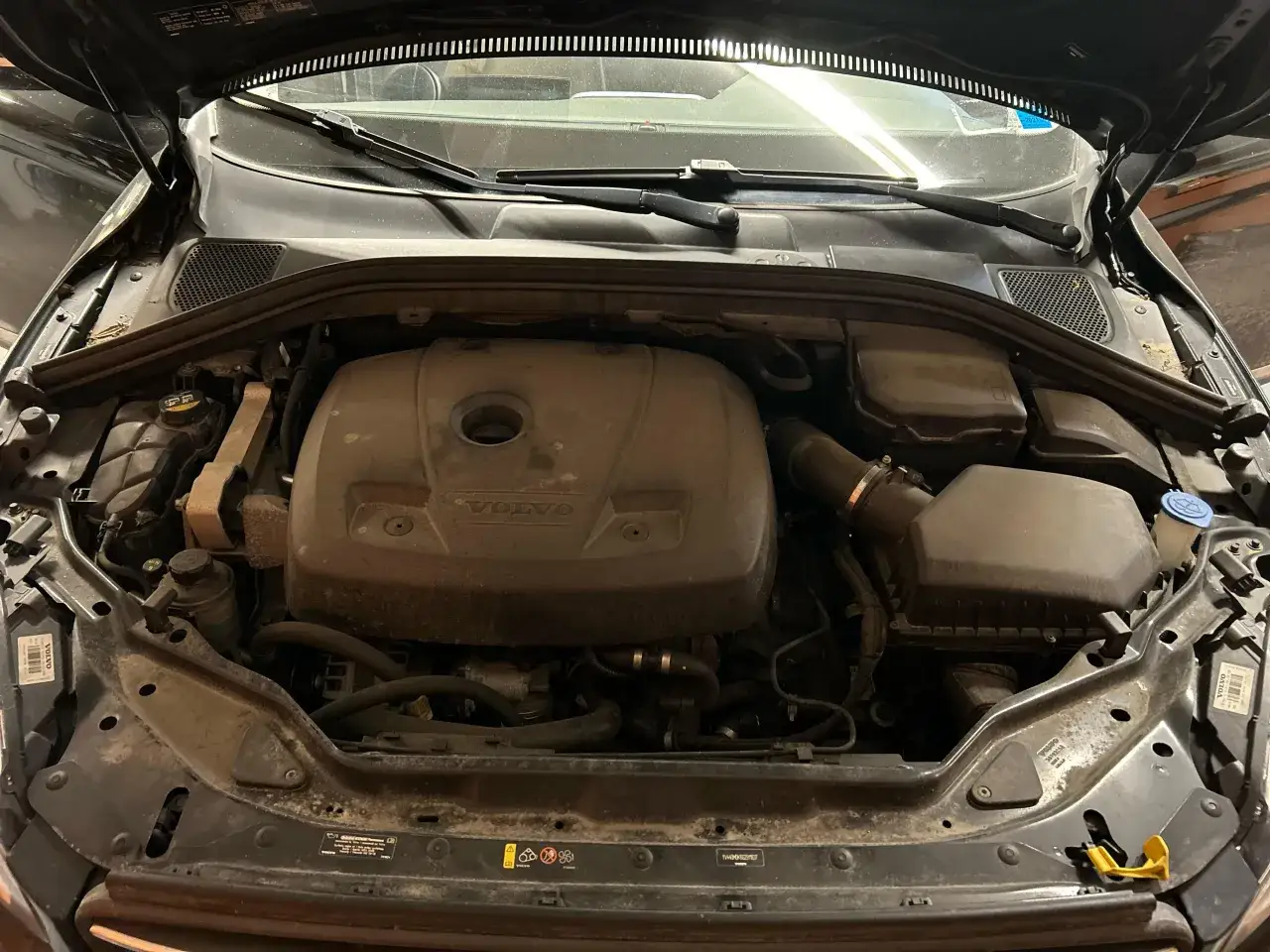 Silnik Volvo S60, widoczny pod maską, jest zakurzony. Widać pokrywę silnika z logo Volvo, elementy układu dolotowego i zbiornik płynu. Akumulator nie jest widoczny.
